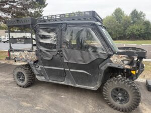 A Can-Am Defender Max with OEM half doors and framed upper soft doors is parked on asphalt near grass, trees, and a fence; it features camo accents, a roof rack, and large all-terrain tires.