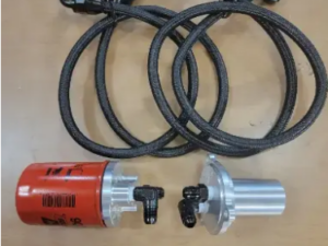 The Can-Am Defender Remote Oil Filter Kit includes two braided black hoses with metal fittings, a red cylindrical filter, and a silver component with black fittings, all neatly arranged on a light brown surface.