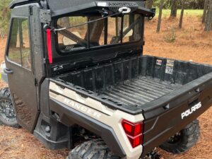 A black and tan Defender Game Loader utility vehicle with rugged tires and a metal roof rack is parked on a pine needle-covered forest floor. Its rear cargo bed is empty, and green-needled pine trees surround the area.