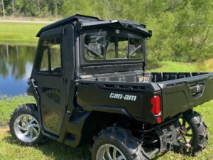 A black Can-Am Defender Game Loader side-by-side utility vehicle with large off-road tires, chrome rims, and a closed cab is parked on grass near a pond, with lush green trees and blue sky in the background.